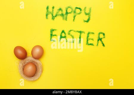 Inschrift Happy easter aus Gras mit Haseneiern und Nest auf gelbem Hintergrund. Platz für Text kopieren Stockfoto
