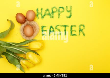 Inschrift Happy easter aus Gras mit Haseneiern, Blumen Tulpen und Nest auf gelbem Hintergrund. Platz für Text kopieren Stockfoto