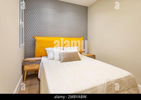 Das Innere des Schlafzimmers mit Bett in der Mitte ist gefüllt mit cremefarbenen Jacquard Bettüberwürfen. Die Kissen sind ordentlich auf dem gelben Kopfteil an der grauen Wand gestapelt Stockfoto