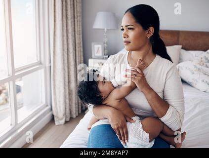 Das wird jeden Tag zu einem größeren Kampf. Eine attraktive junge Frau, die zu Hause auf dem Bett sitzt und ihre kleine Tochter mit einer Flasche füttert. Stockfoto