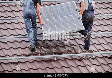 Männer, die Photovoltaik-Solarmodule auf dem Dach des Hauses tragen. Schnittbild von Elektrikern, die Solarpaneelsysteme im Freien installieren. Konzept alternativer und erneuerbarer Energien. Stockfoto