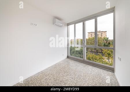 Geräumiges, leeres Zimmer mit weißen Wänden, raumhohen Fenstern und Marmorboden. An der Wand spart die Klimaanlage die Sommerhitze. Aus dem Fenster des Zimmers Stockfoto