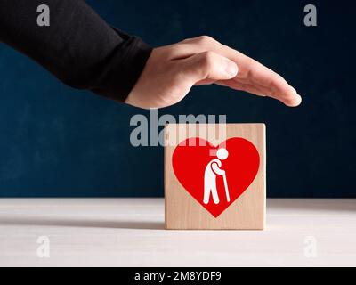 Konzept für Sicherheit, Unterstützung, Versicherung oder Betreuung von Senioren. Männliche Hand steht mit einer Ikone eines älteren Ruheständlers protektiv über dem Holzwürfel. Stockfoto