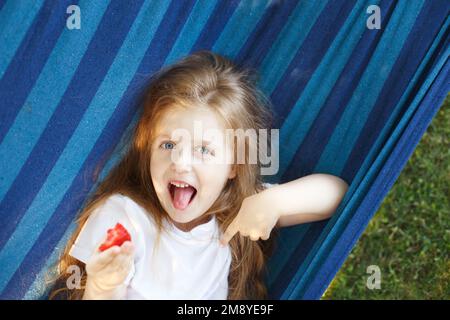 Ein blondes Mädchen mit langen Haaren isst eine Erdbeere im Garten, die in einer Hängematte liegt. Stockfoto