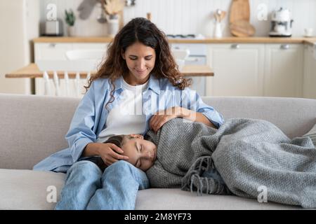 Tired child daughter covered with warm blanket fall asleep after school, sleeping on mother knees Stock Photo