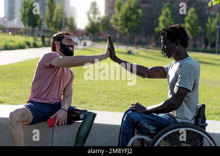 Zwei Freunde, ein Latino-Mann mit Bart und Beinprothese, und ein Afrikaner im Rollstuhl, High-Five-gegenseitig in einer städtischen Umgebung im Freien Stockfoto