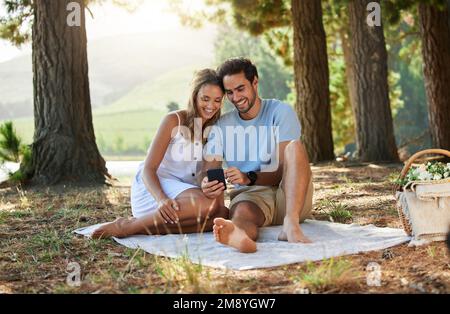 Ich habe gute Neuigkeiten für Sie. Ein Paar, das sich auf einem digitalen Tablet etwas anschaut, während es im Wald ein Picknick macht. Stockfoto