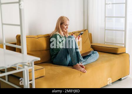 Kaukasische Frau mittleren Alters, die mit dem Handy im Internet surft, während sie zu Hause auf dem gelben Sofa sitzt. Stockfoto