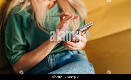 Unerkannte Frau, die auf dem Mobiltelefon in den Medien surft, während sie zu Hause auf dem gelben Sofa sitzt. Stockfoto