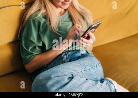 Unerkannte Frau, die auf dem Mobiltelefon in den Medien surft, während sie zu Hause auf dem gelben Sofa sitzt. Stockfoto