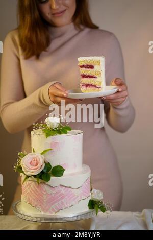 Mädchen schneidet und serviert ein Stück Kuchen. Festliche Hochzeit zweistufigen Kuchen mit frischen Blumen auf einem grauen Hintergrund dekoriert Stockfoto