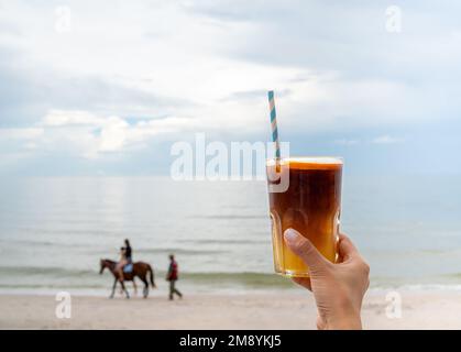 Ein Glas frisch gefrorenes orangefarbenes Ameriano, dekoriert mit geschnittenem ​​orange, das von Hand auf dem Strandhintergrund gehalten wird, mit Blick auf das Meer. Kalter schwarzer Kaffee mit o Stockfoto