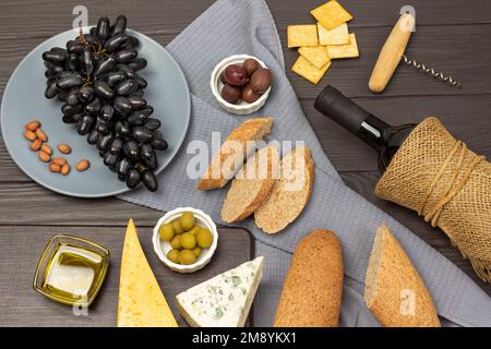 Baguette und Oliven in Schüsseln auf einer grauen Serviette. Schwarze Trauben auf einem grauen Teller. Käse, eine Flasche Wein und Cracker. Flach verlegt. Dunkler Holzhintergrund. Stockfoto