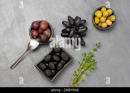 Grüne, rote und schwarze Oliven in Schüsseln. Thymianzweig und ein Haufen schwarze Trauben. Speicherplatz kopieren. Flach verlegt. Grauer Hintergrund. Stockfoto
