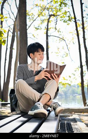 Porträt, gutaussehender und schauriger junger asiatischer Student, der ein Buch liest, während er sich am Wochenende im Park entspannt. Das Leben von Universitätsstudenten Stockfoto