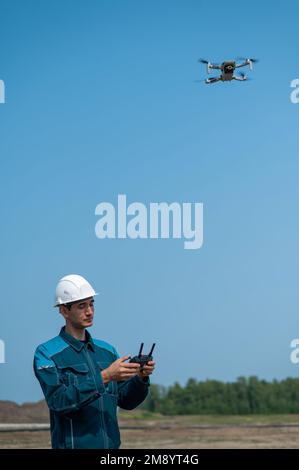 Ein Mann in Helm und Overall steuert eine Drohne auf einer Baustelle. Der Bauherr führt die technische Überwachung durch. Stockfoto