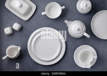 Weißes Geschirr zum Servieren und Essen. Leere saubere Teller, Tasse, Teekanne, Zuckerdose, Milchkanne und Butterschale auf einem grauen Betontisch, Draufsicht Stockfoto