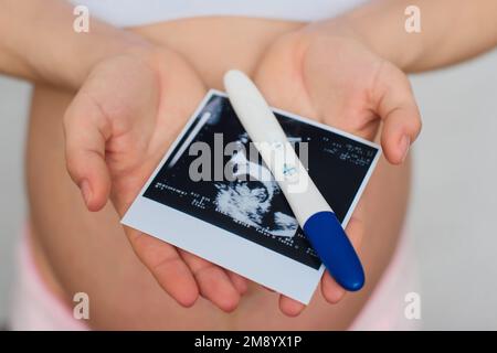 Ultraschallbild und positiver Schwangerschaftstest in den Händen eines schwangeren Mädchens am Hintergrund des Abdomens Stockfoto