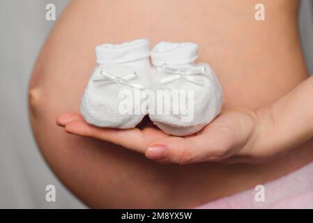 Neugeborenenstiefel in den Händen eines schwangeren Mädchens am Hintergrund des Abdomens Stockfoto