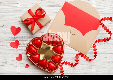 Umschlag auf farbigem Hintergrund zum Valentinstag mit Geschenkbox und Schokolade. Herzförmig mit Geschenkschachtel mit Pralinen Draufsicht mit Kopierraum. Stockfoto