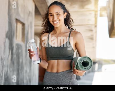 Bereit für das Workout. Eine attraktive junge Frau steht allein und hält eine Wasserflasche und Yoga-Matte während ihres Workouts im Freien. Stockfoto