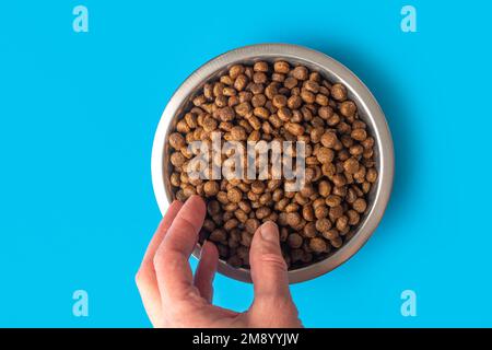 Eine Hand, die trockenes Haustierfutter in einer Metallschüssel auf blauem Hintergrund aufträgt. Fütterung von Katzen und Hunden mit granuliertem Trockenfutter. Pellets mit gesundem Futter für Hunde und Stockfoto