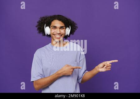Junger, fröhlicher, attraktiver lateinamerikanischer Lockenmann mit Kopfhörern, der zur Seite zeigt und in die Kamera schaut, während er über einem isolierten violetten Hintergrund steht Stockfoto