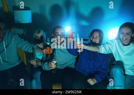 Freunde, die im Wohnzimmer fernsehen, Bier trinken, fröhlich schreien, abends zusammen im Fernsehen Sport sehen. Stockfoto