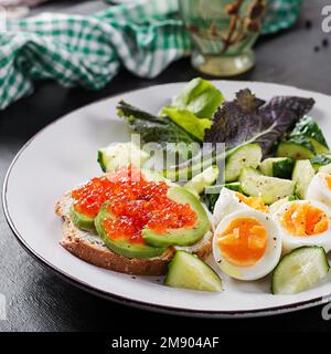 Frühstück. Gesundes Sandwich auf Toast mit Avocado und rotem Kaviar, gekochten Eiern, Gurkensalat auf weißem Teller. Gesunde Proteinkost. Stockfoto