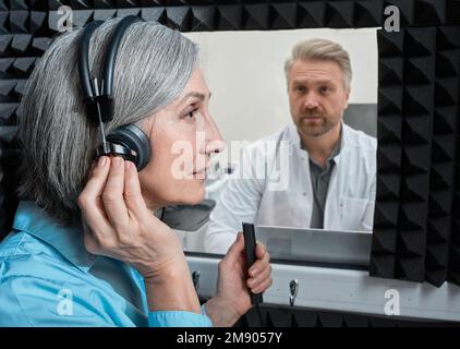 Hörtest. Patientin mit audiologischen Kopfhörern drückt auf Tastendruck während der audiometrischen Prüfung in einer schalldichten Audiologiekabine Stockfoto