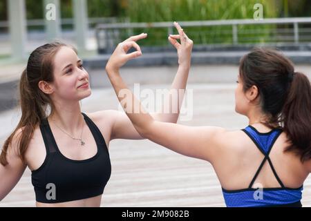 Zwei fröhlich lächelnde, wunderschöne weibliche Teenager-Freunde üben am Wochenende Yoga-Übungen im Stadtpark im Freien. Gesunder Bewegungssport und Lifest Stockfoto