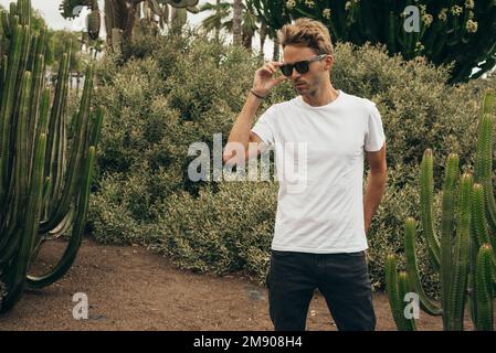 Ein blonder, attraktiver Hipstertyp mit weißem, leerem Rührarm und Sonnenbrille steht im Garten neben einem großen Kaktus. Stockfoto