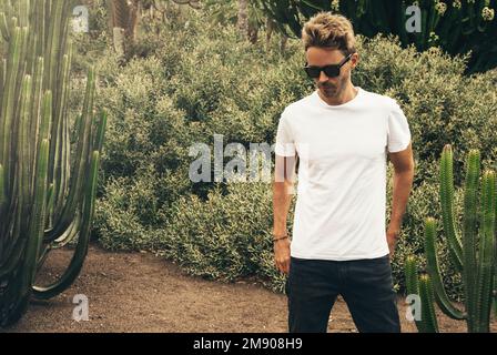 Ein blonder, attraktiver Hipstertyp mit weißem, leerem Rührarm und Sonnenbrille steht im Garten neben einem großen Kaktus. Stockfoto