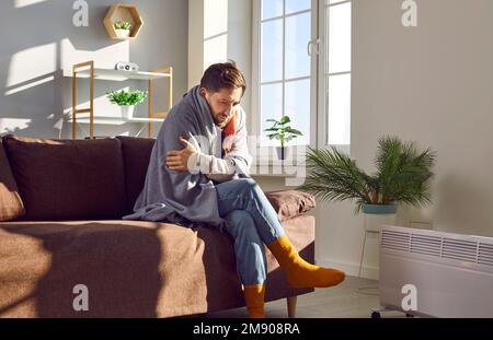 Ein junger Mann mit einer Decke, der auf der Couch neben der Heizung saß und versuchte, sich aufzuwärmen Stockfoto