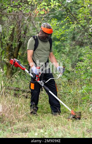 Junger erwachsener Mann in Schutzkleidung, der im Garten grünes Gras mit einem elektrischen Rasentrimmer mäht. Seitenansicht. Stockfoto