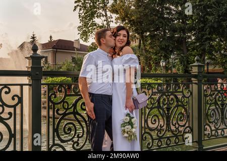 Der junge Mann küsst sanft seine Freundin und umarmt sie auf einer Brücke in der Nähe eines Teichs mit Springbrunnen. Ein Mädchen mit Blumen lächelt glücklich. Stockfoto