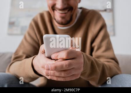 Zugeschnittene Ansicht eines glücklichen Mannes in beigefarbenem Pullover mit Smartphone zu Hause, Stockbild Stockfoto