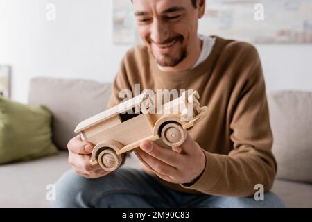 Ein bärtiger Mann lächelt, während er im Wohnzimmer auf ein hölzernes Autospielzeug schaut, Stockbild Stockfoto