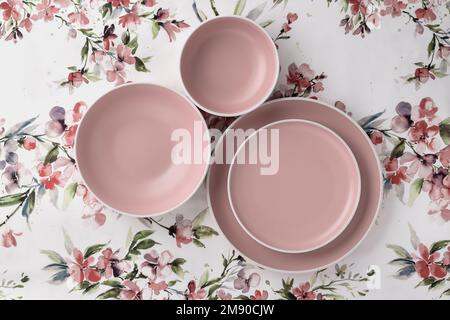 Leere saubere Teller auf einer weißen Tischdecke mit Blumenmuster, Blick von der Tischplatte. Flach liegend, minimalistisches Design. Rosafarbenes Keramikgeschirr. Trendiges Geschirr Stockfoto