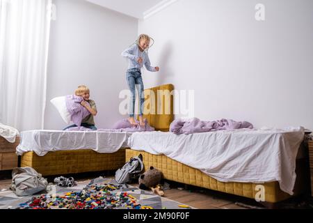 Geschwister, die Spaß haben, lachen, Junge und Mädchen spielen Kissenschlacht in einem dreckigen Kinderzimmer, Kinder spielen unter den vielen Spielzeugen zu Hause, viele Spielzeuge und Stockfoto