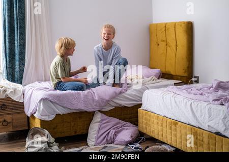 Geschwister, die Spaß haben, lachen, Junge und Mädchen spielen Kissenschlacht in einem dreckigen Kinderzimmer, Kinder spielen unter den vielen Spielzeugen zu Hause, viele Spielzeuge und Stockfoto