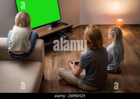 Drei Kinder spielen das Videospiel Green Screen Chromakey auf der tv-Konsole mit dem Joystick, während sie zu Hause sitzen, Brüder und Schwestern sitzen auf dem Boden Stockfoto