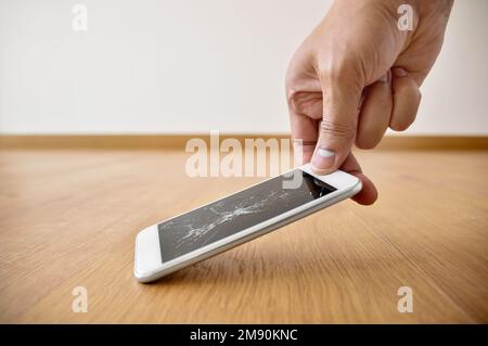 Person, die zu Hause ein kaputtes Smartphone mit einem rissigen Bildschirm auf dem Boden aussucht Stockfoto