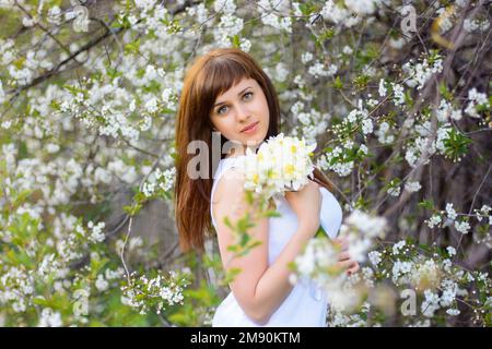 Hübsches junges Mädchen mit einem Strauß Narzissen in einem weißen Kleid auf einem Hintergrund von Kirschblüten im Frühling Stockfoto