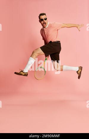 Porträts eines gutaussehenden charismatischen Mannes in stilvoller Kleidung mit Tennisschläger in einem Sprung über pinkfarbenen Studiohintergrund. Das Konzept von Mode, Sport Stockfoto