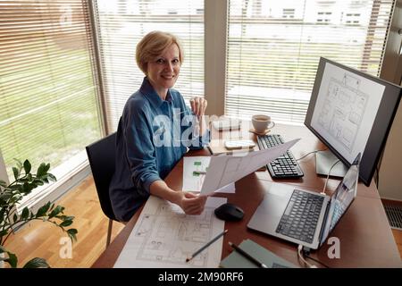 Eine lächelnde, reife Ingenieurin arbeitet vom Heimbüro aus mit einem Computer an einem Projekt Stockfoto