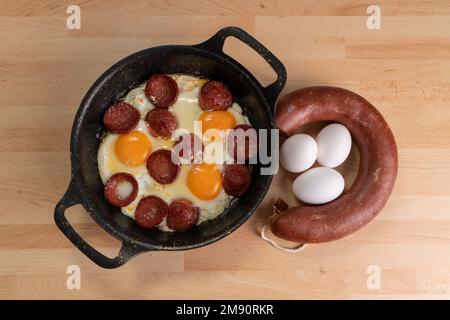 Blick von oben auf Spiegeleier mit Speckscheiben. Rohe Wurst und unzerbrochene Eier befinden sich neben der Pfanne auf Holzhintergrund. Stockfoto