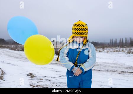 Porträt eines traurigen, unglücklichen Jungen, der allein auf dem schneebedeckten Feld steht, mit gelben blauen Ballons. Auf Krieg, militärische Angriffe und Terror in der Ukraine aufmerksam machen Stockfoto