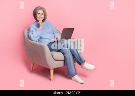 Ganzkörpergröße Kaderoptimistin Rentnerin Frau sitzt weicher Stuhl Laptop modernes Gerät Touch Kinn für geschäftliche Aufgaben isoliert auf pinkfarbenem Hintergrund Stockfoto