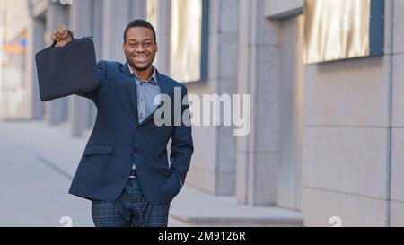 Fröhlich lächelnder männlicher afroamerikanischer Geschäftsmann, ethnischer Arbeiter, Unternehmer auf der Stadtstraße, hält und zeigt Aktentaschen Stockfoto